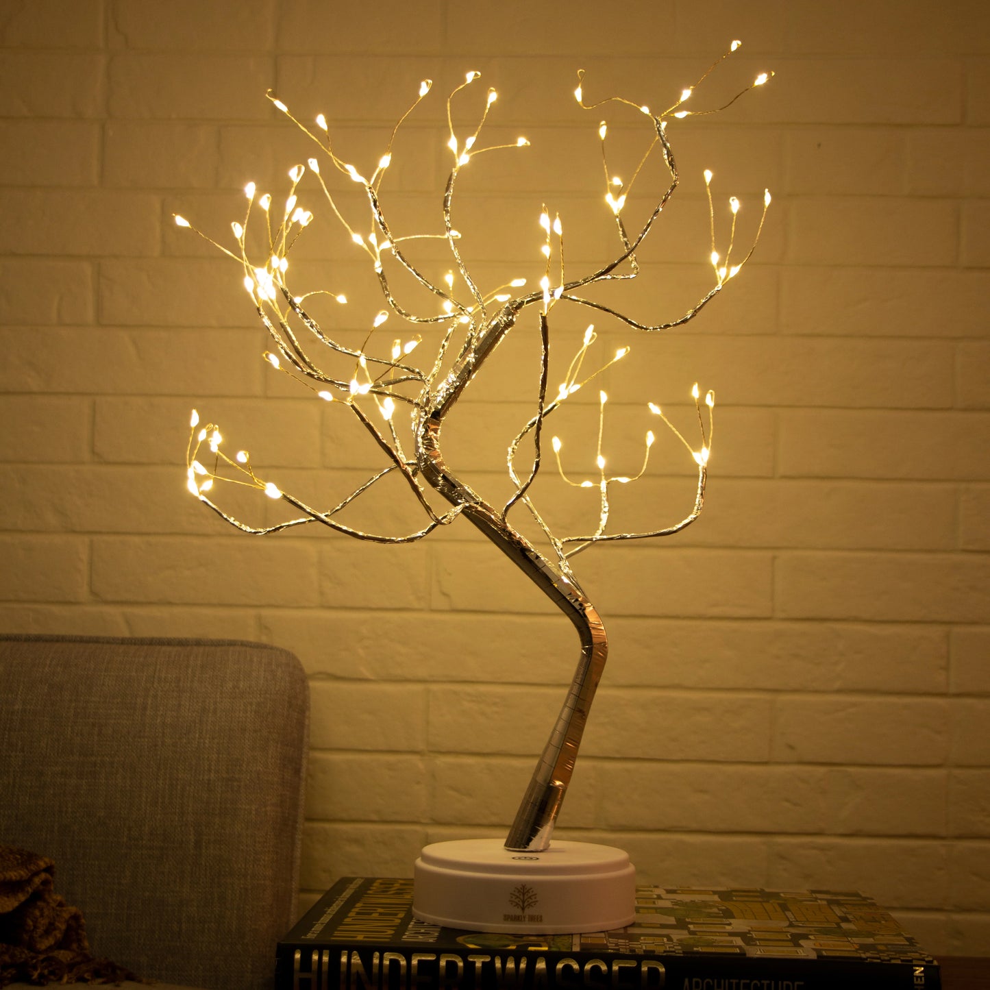 Decorative LED tree with lights on a stand against a brick wall.
