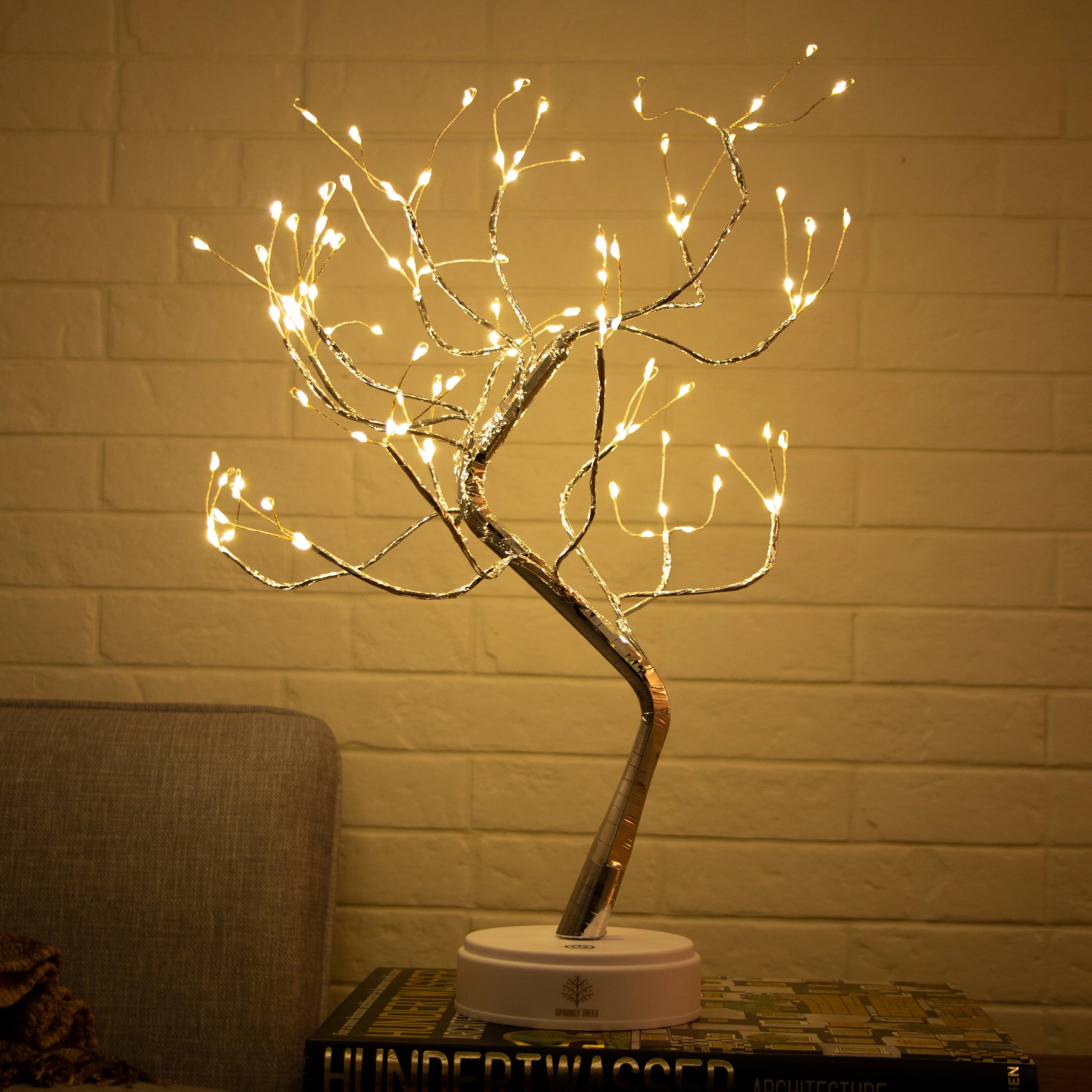 Decorative LED tree with lights on a stand against a brick wall.