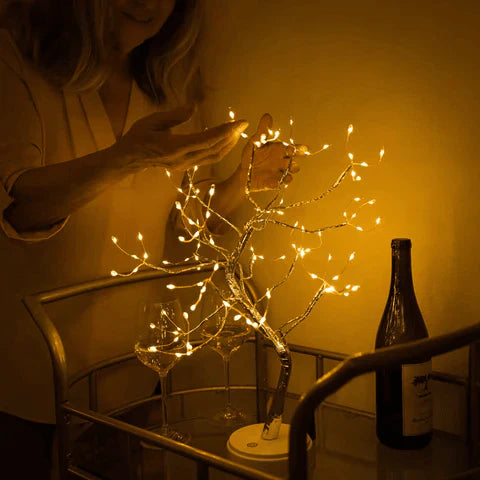 Person holding a small tree with lights on a table next to a bottle of wine.